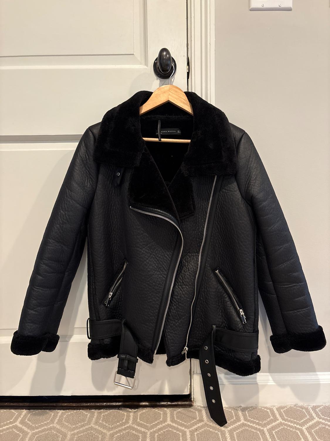 Black textured jacket with zips, belt and faux fur lining. ZARA WOMAN label. Hanging on a wooden hanger on a white door.
