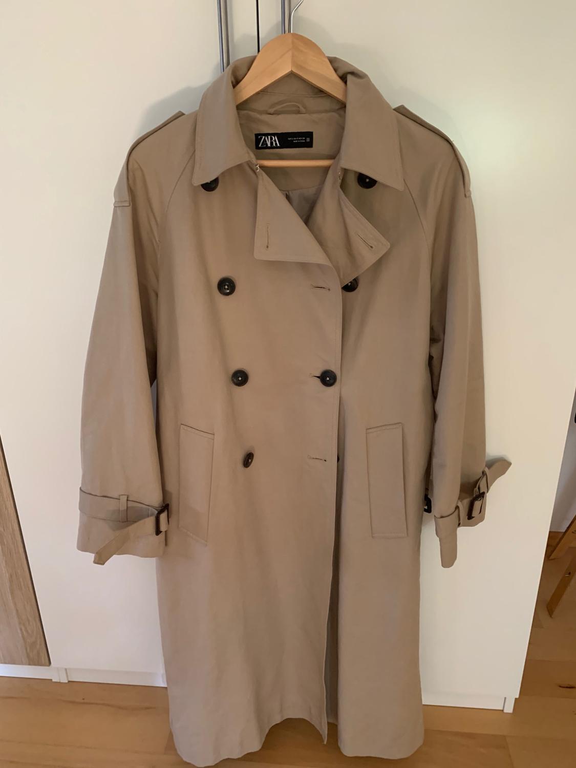 Beige trench coat with dark buttons hanging on a wooden hanger against a white background. The text ZARA can be read on the inner label.