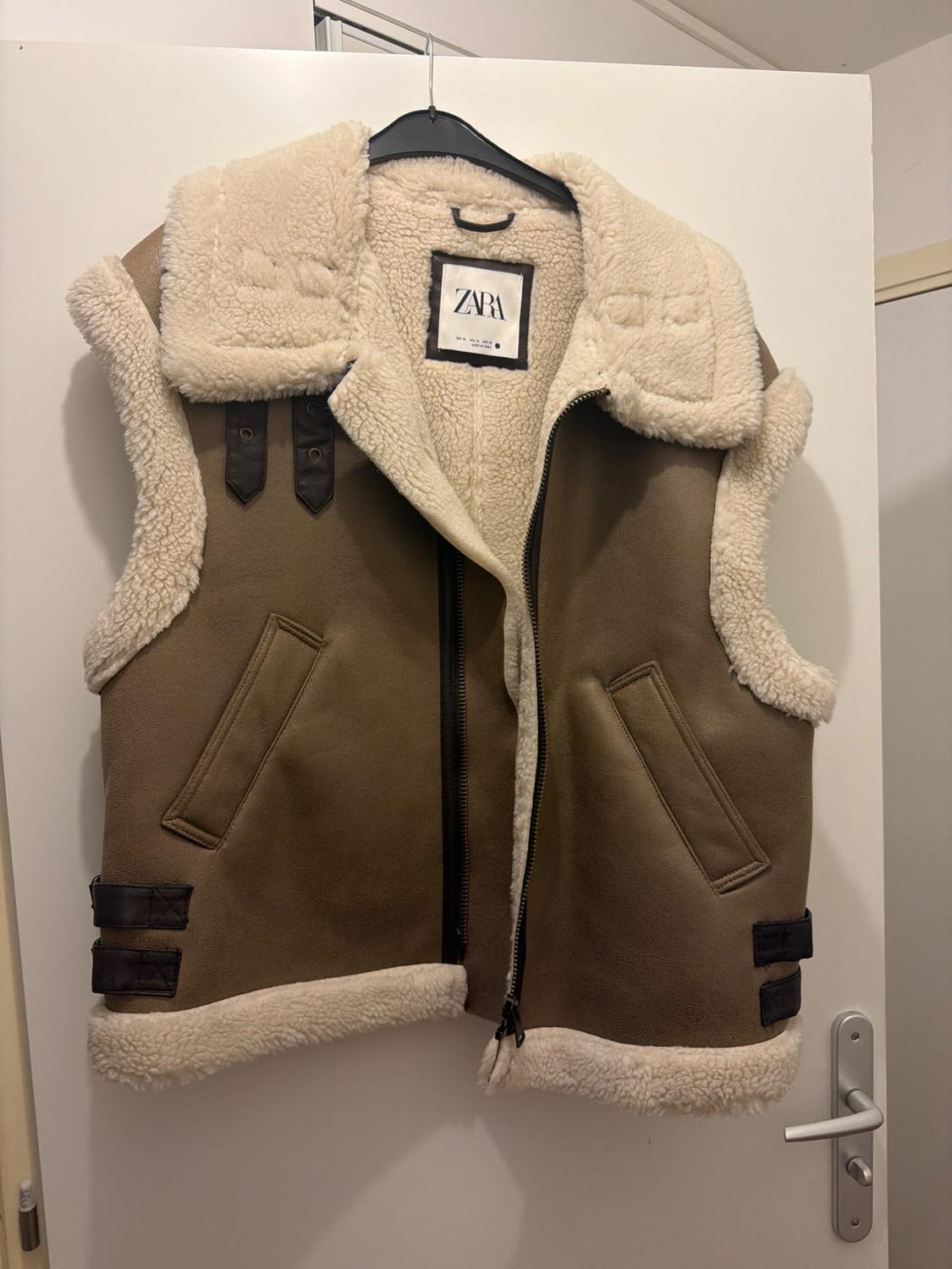 Brown gilet with a light fur lining and zip, hanging on a hanger over a white door. Inner label bearing the text ZARA.