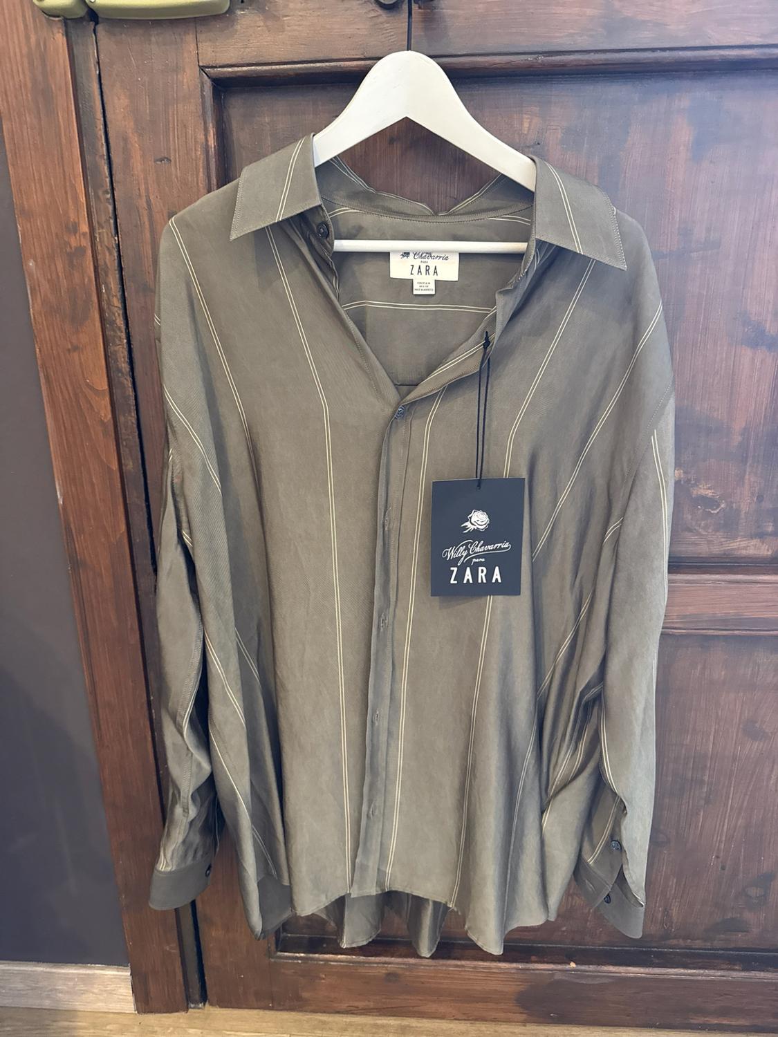 Green long-sleeved shirt with white stripes hanging on a wooden door. Label bearing the text Willy Chavarria for ZARA.