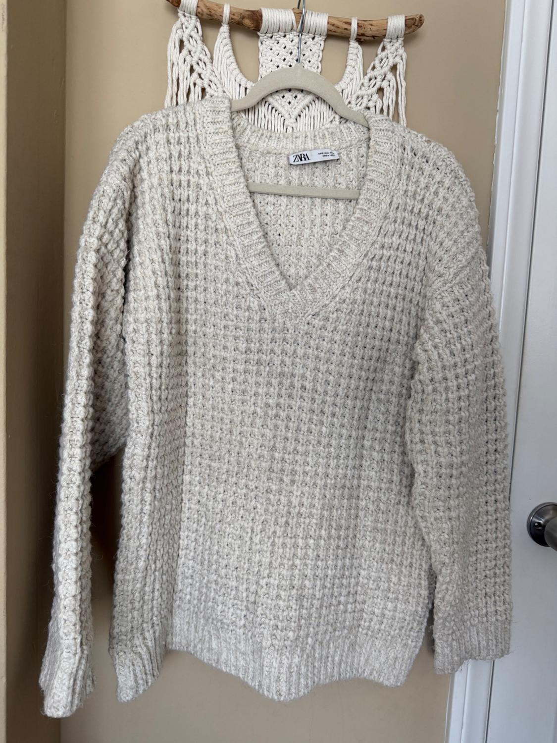 Chunky cream knit sweater hanging on a hanger against a macrame background. Visible tag with the ZARA brand.