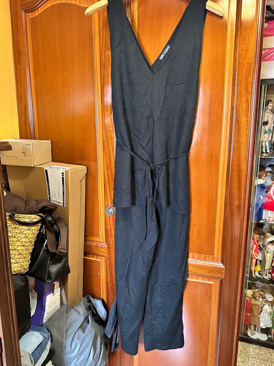 Black jumpsuit hanging on a wooden door, surrounded by boxes and bags. In the background, a display cabinet with dolls.