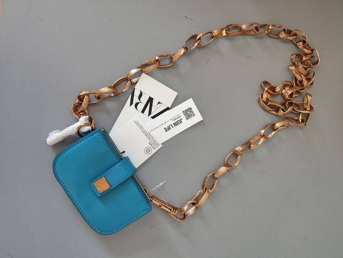 Blue bag with gold chain on a grey background. Labels with the text ZARA, JOIN LIFE, LEATHER, OUTER SHELL, CARE FOR PLANET.