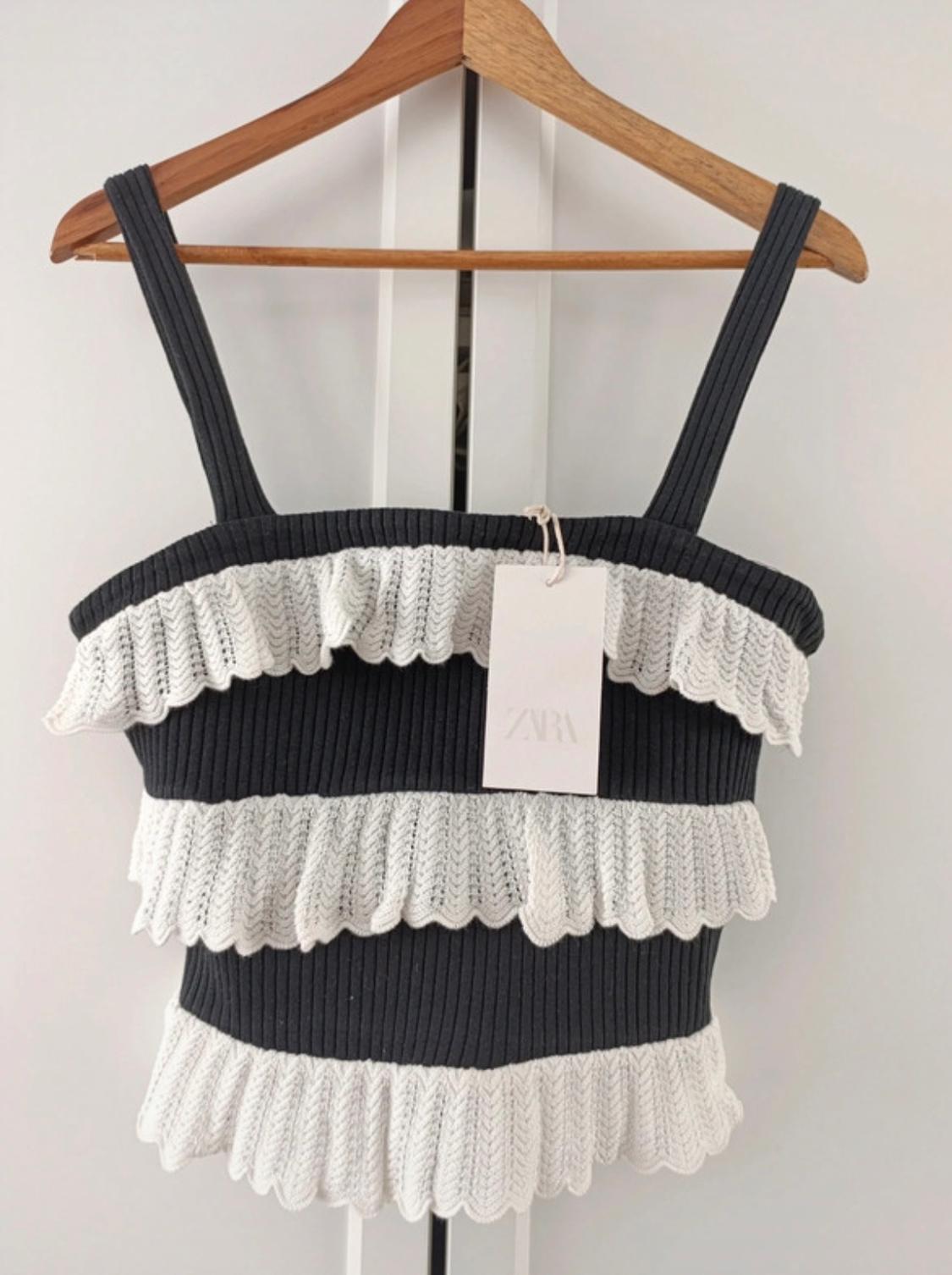 Black camisole with white ruffles on a wooden hanger. Visible label with text "ZARA". White background.