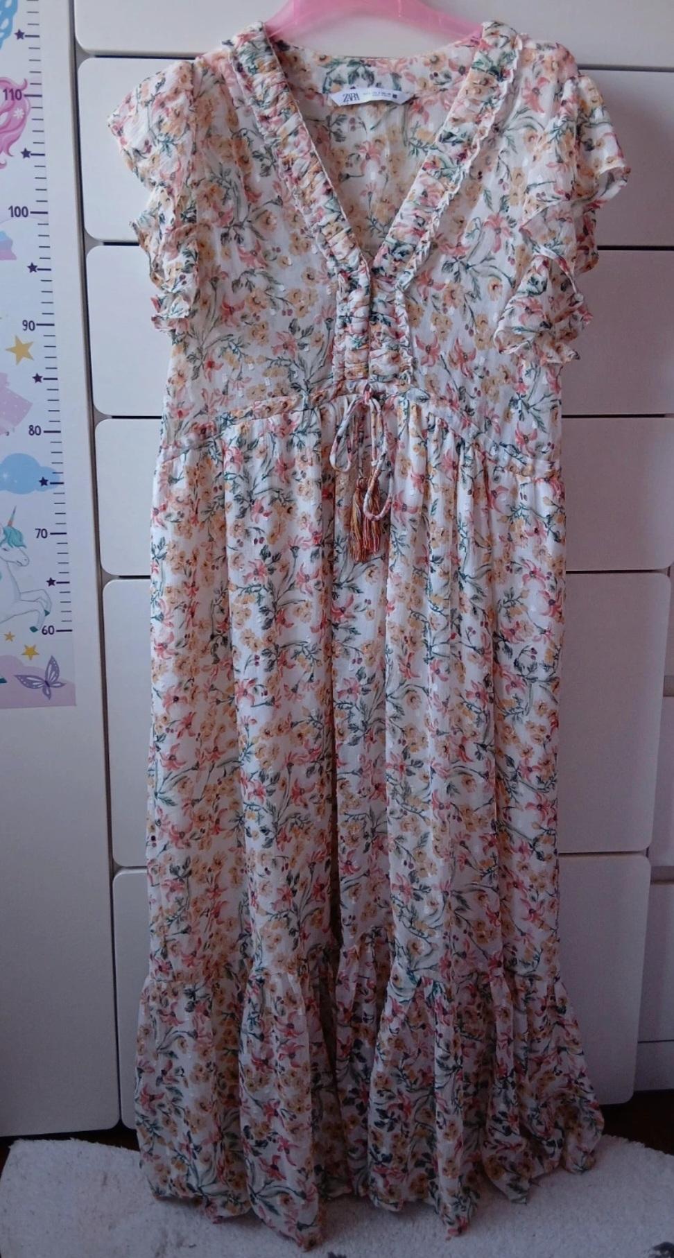 Floral maxi dress with ruffles and V-neck, hanging on a pink hanger. Background of white drawers and child's height chart.