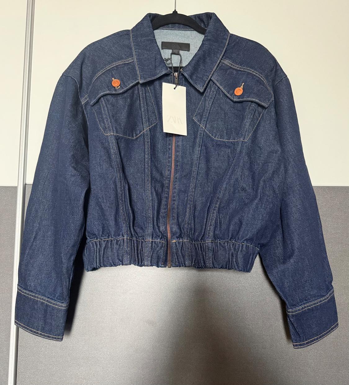 Dark blue denim jacket with a zip, elasticated hem and pockets, hanging on a hanger. It has a label with the text ZARA.