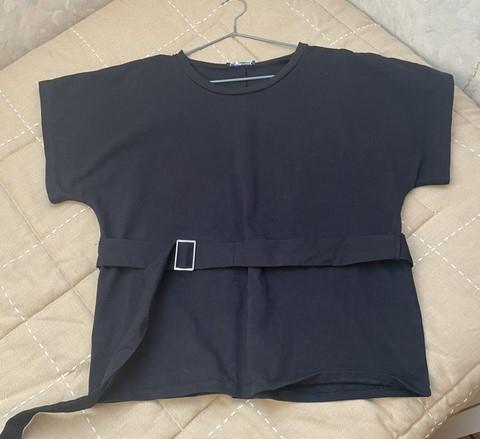 Black short-sleeved T-shirt with adjustable belt and square buckle, hanging on a hanger over a beige padded surface.