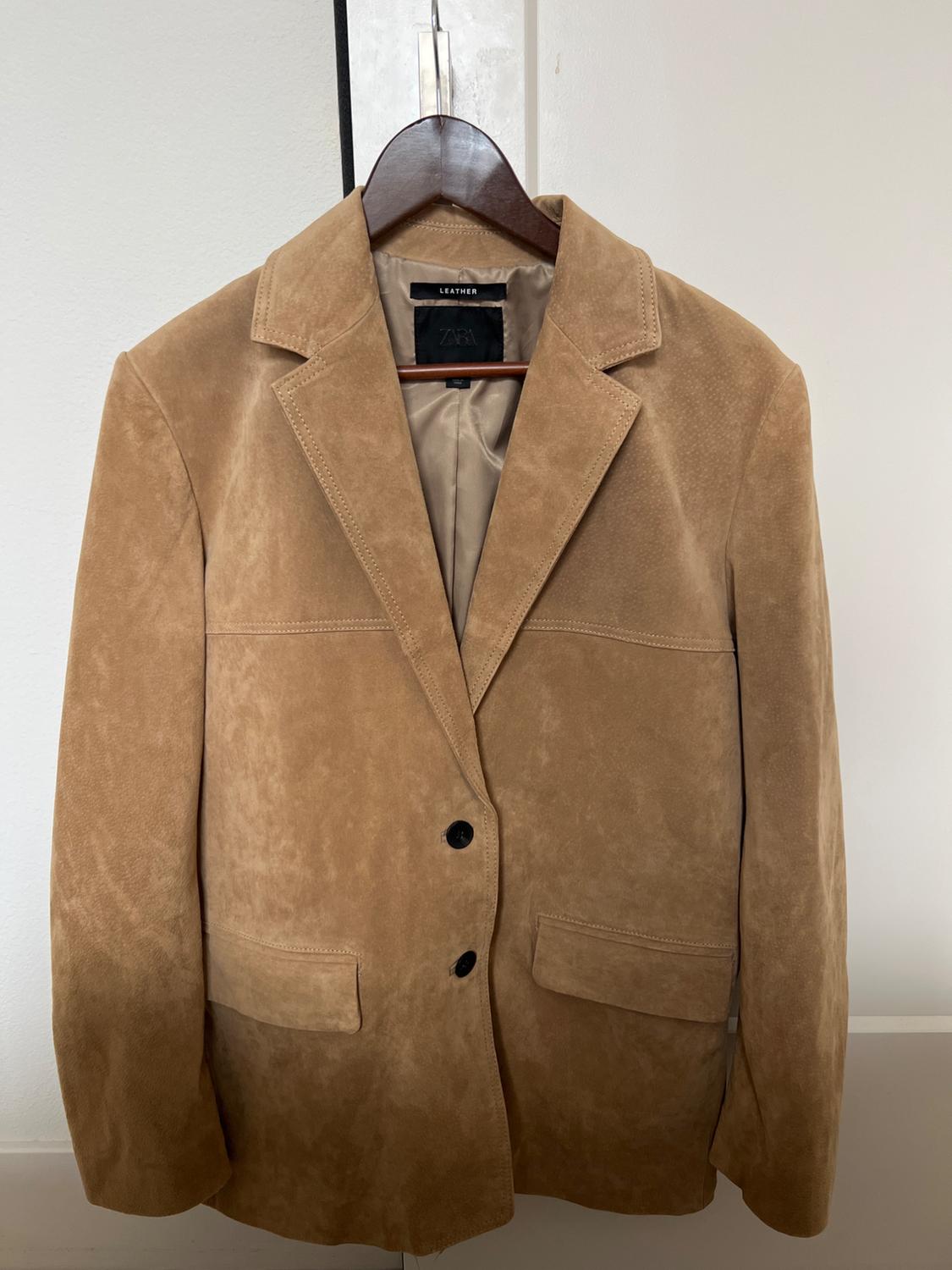 Light brown leather jacket hanging on a wooden hanger. Visible tag: "ZARA LEATHER". Plain white background.