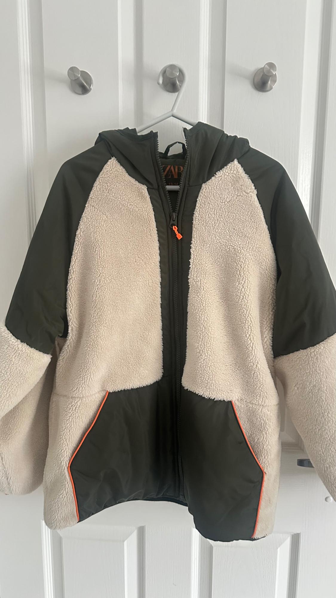 ZARA hooded jacket hanging on a white door. It combines dark green fabric
and beige fleece material with orange details.