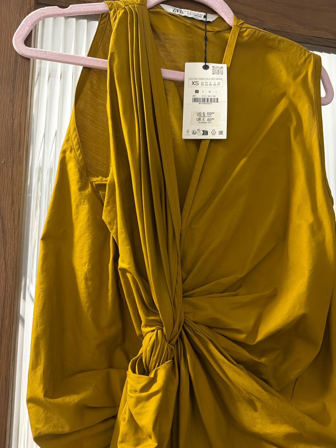Mustard yellow dress with front draping and a price tag hanging, suspended on a pink hanger in front of a mirror.