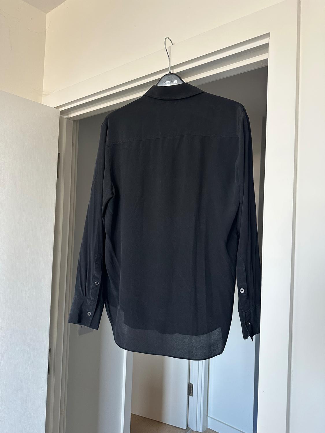 Back of a black long-sleeved shirt on a hanger with the text REISS, hanging on the frame of a white door.