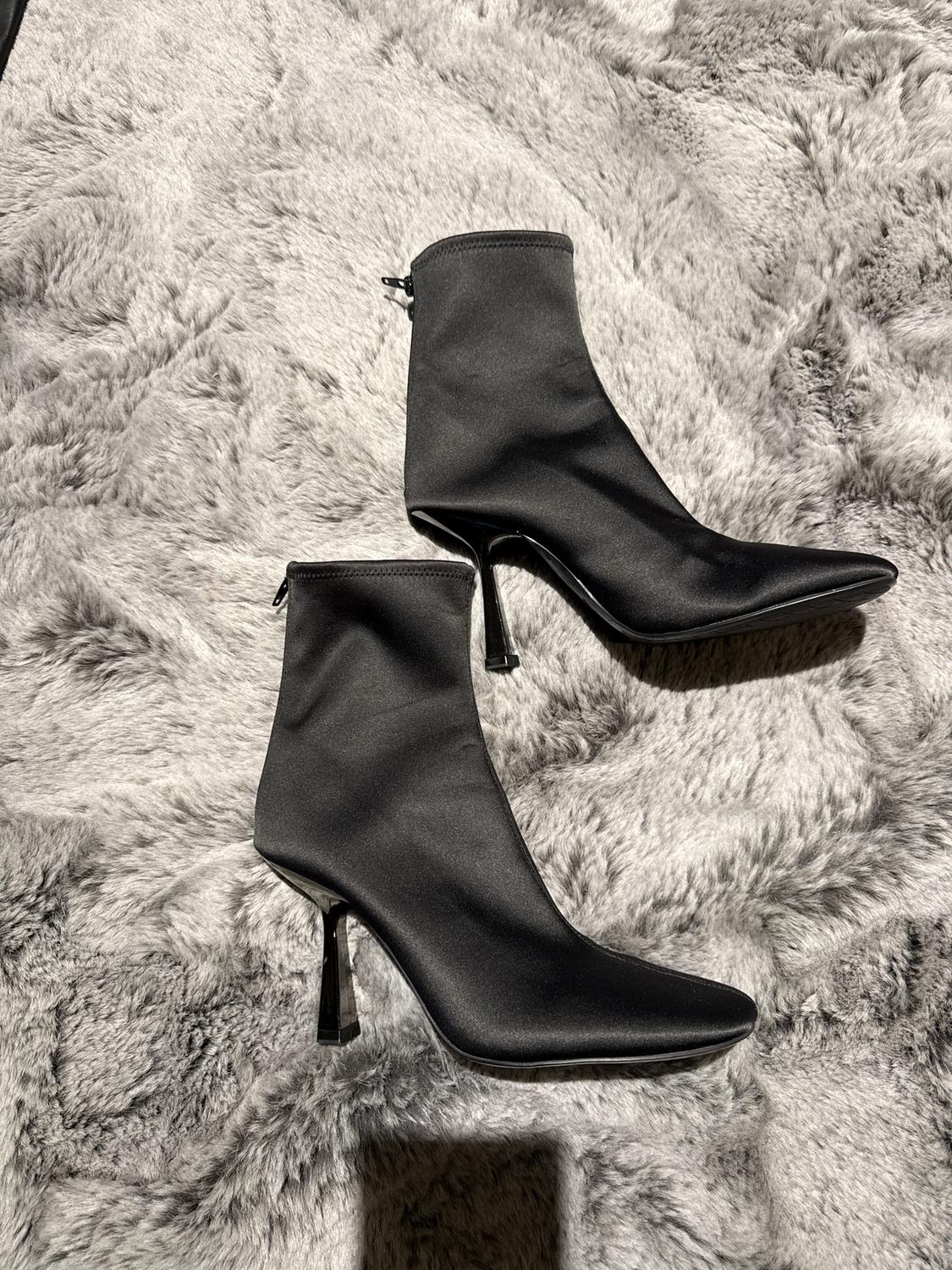 Black high-heeled ankle boots on a fluffy gray rug, illuminated by a soft light that highlights their satin texture.