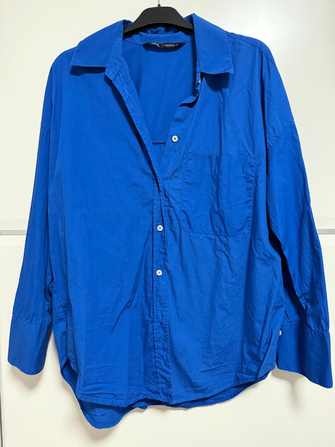 Blue long-sleeved shirt hanging on a black hanger. It has a chest pocket and white buttons. White background.