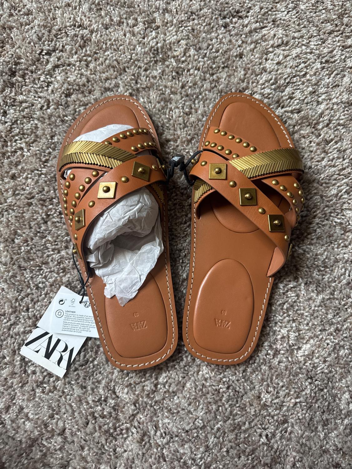 Top view of brown flat sandals with crisscross straps and gold studs on a carpet. Visible text: ZARA, LEATHER, 39.