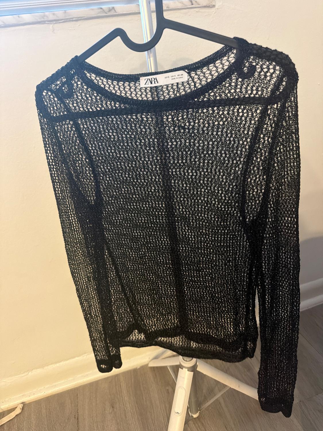 Black open-knit top on a hanger. Text: ZARA EUR S USA S MEX 26 MADE IN CHINA. Relaxed setting with a white wall and wooden floor.