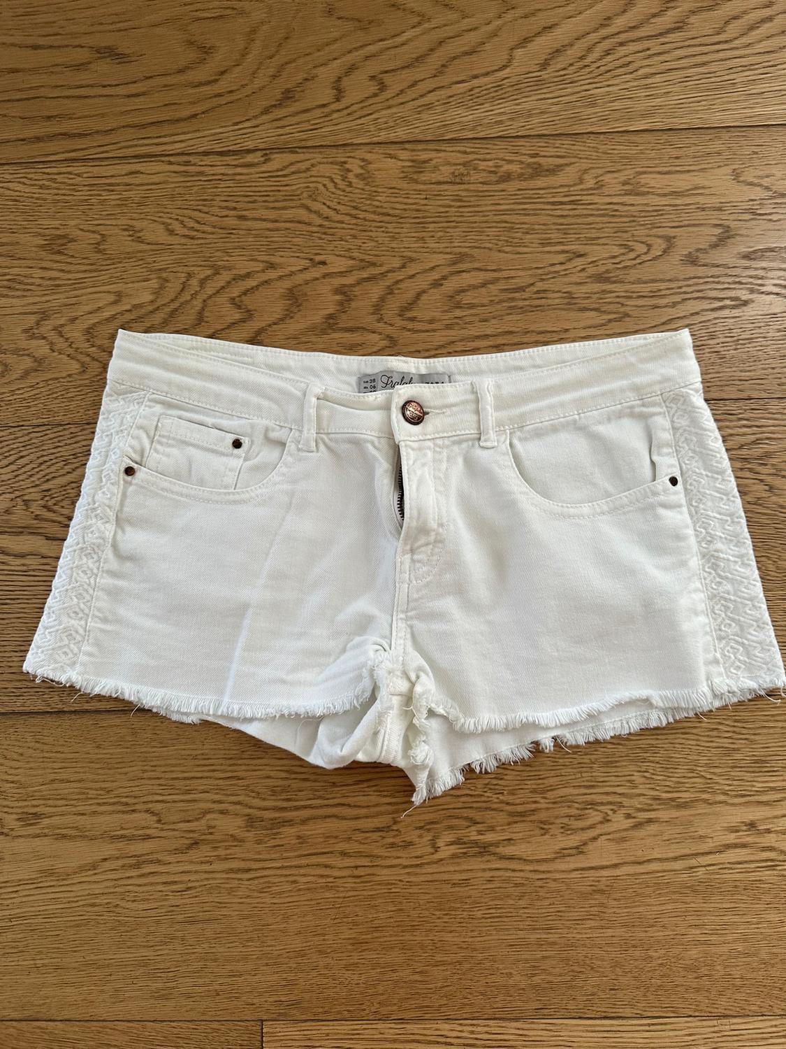 White denim shorts with frayed edges and embroidered details on the sides, on a light wooden floor.