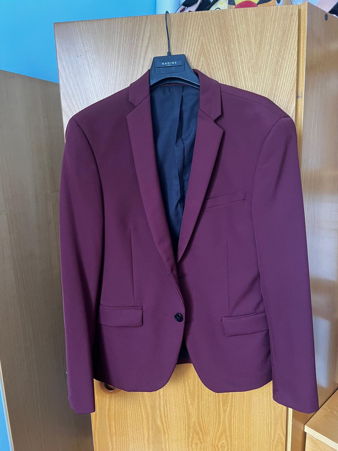 Burgundy jacket hanging on a black hanger with the brand "NADINE", against a light wood background.