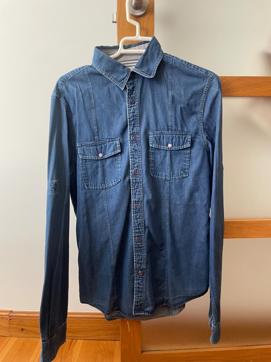 Blue long-sleeved denim shirt with two chest pockets, hanging on a white hanger on a wooden structure against a white background.