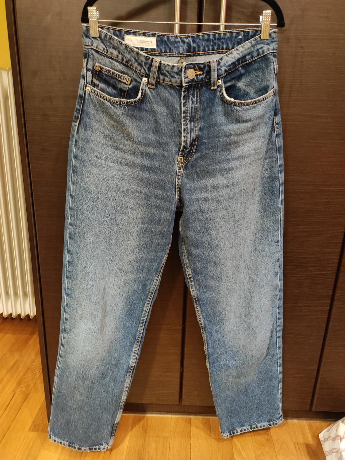 Straight-cut blue jeans hanging on a hanger against a dark wooden background. Light wooden floor visible.