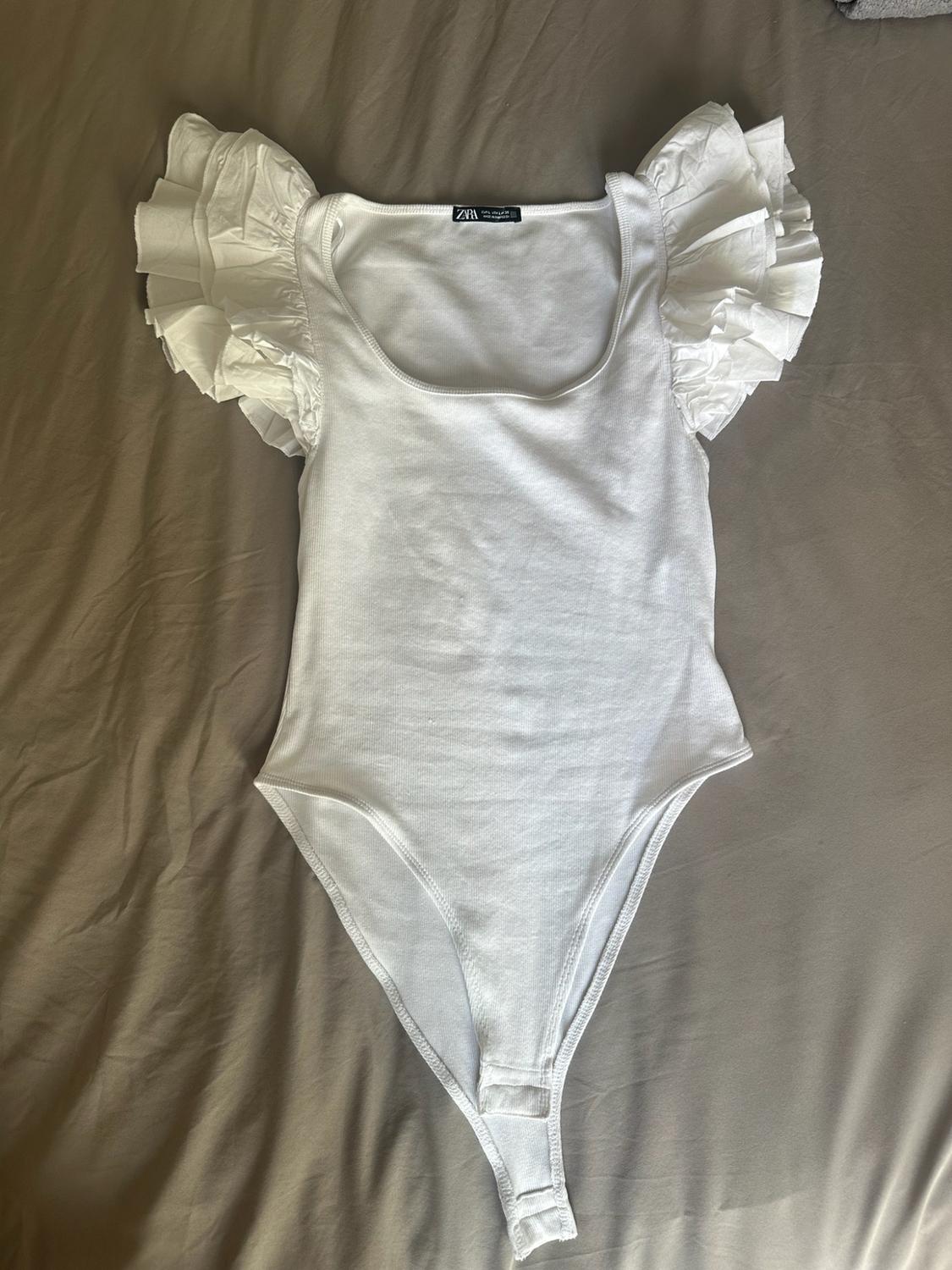 White bodysuit with round neckline and frill sleeves, ZARA label, laid out on a beige sheet.