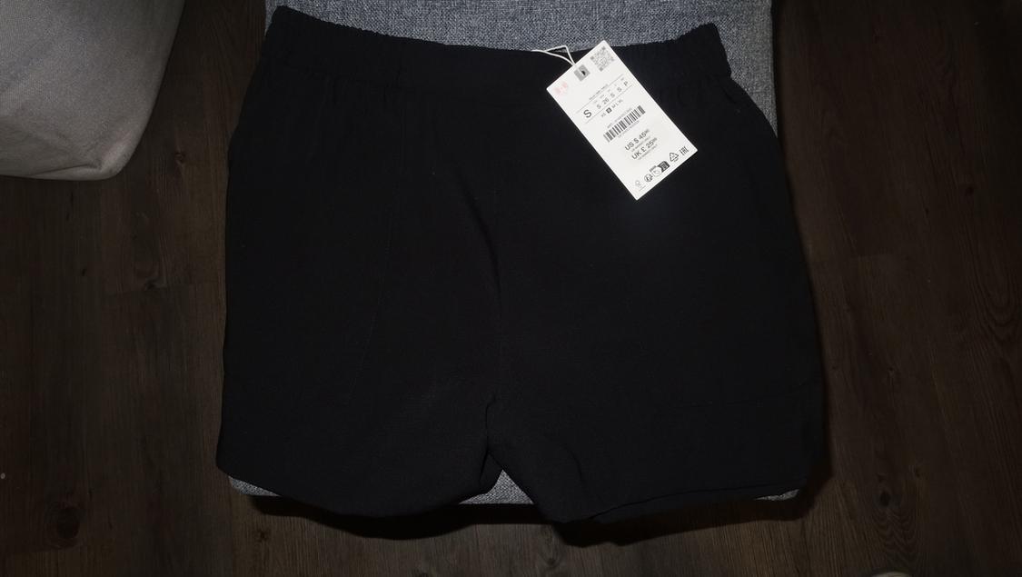 Black shorts on a gray chair, with a visible white tag showing size S and a barcode. Dark wood background.