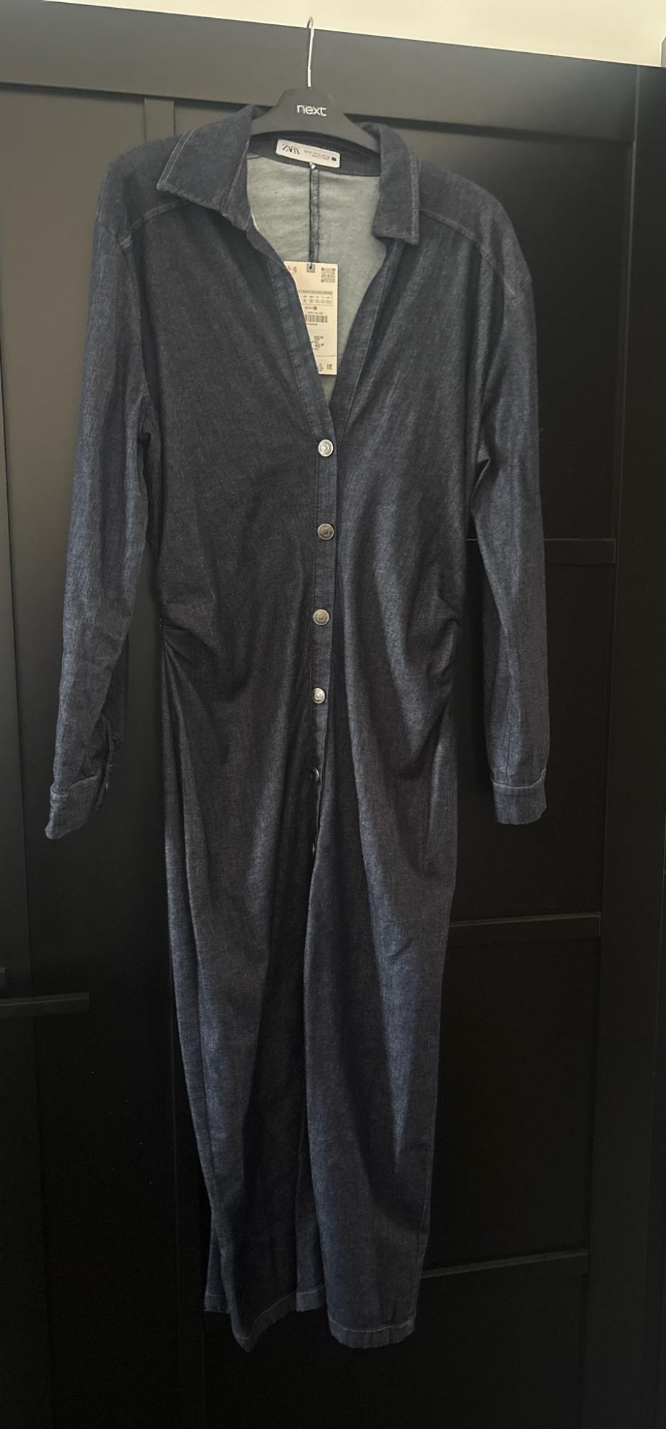 Dark blue long-sleeved dress with front buttons, hanging on a hanger with "next" text on a black door. "ZARA" label visible.