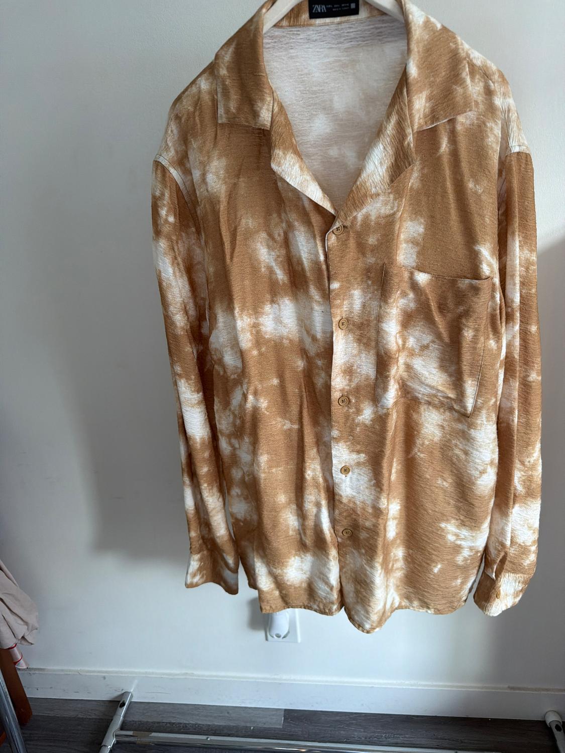 Long-sleeved shirt with a tie-dye print in brown and white tones, hanging on a hanger against a white wall background.