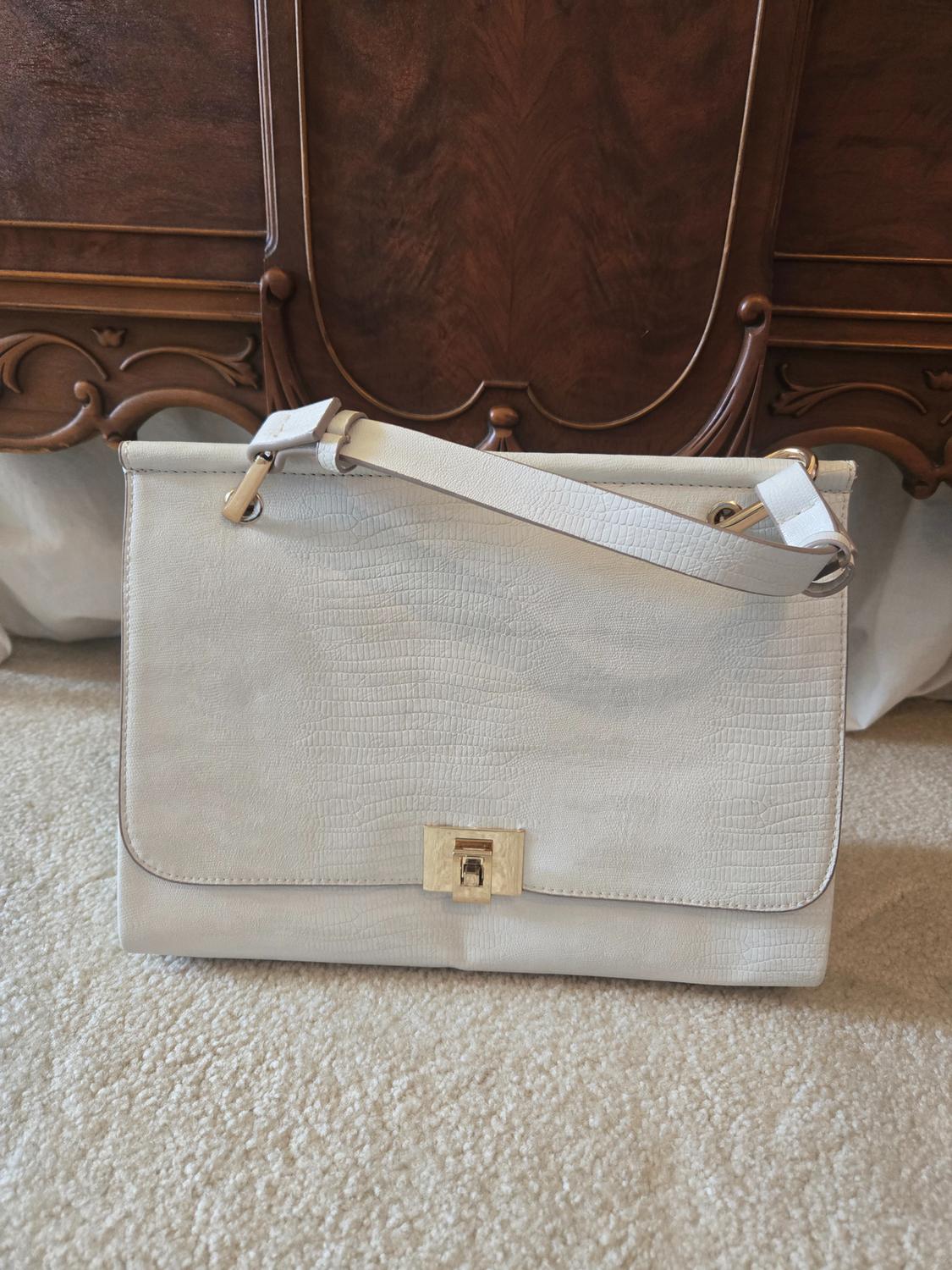 Beige handbag with a leather texture and gold clasp, on a light carpet, in front of a dark wooden cabinet.