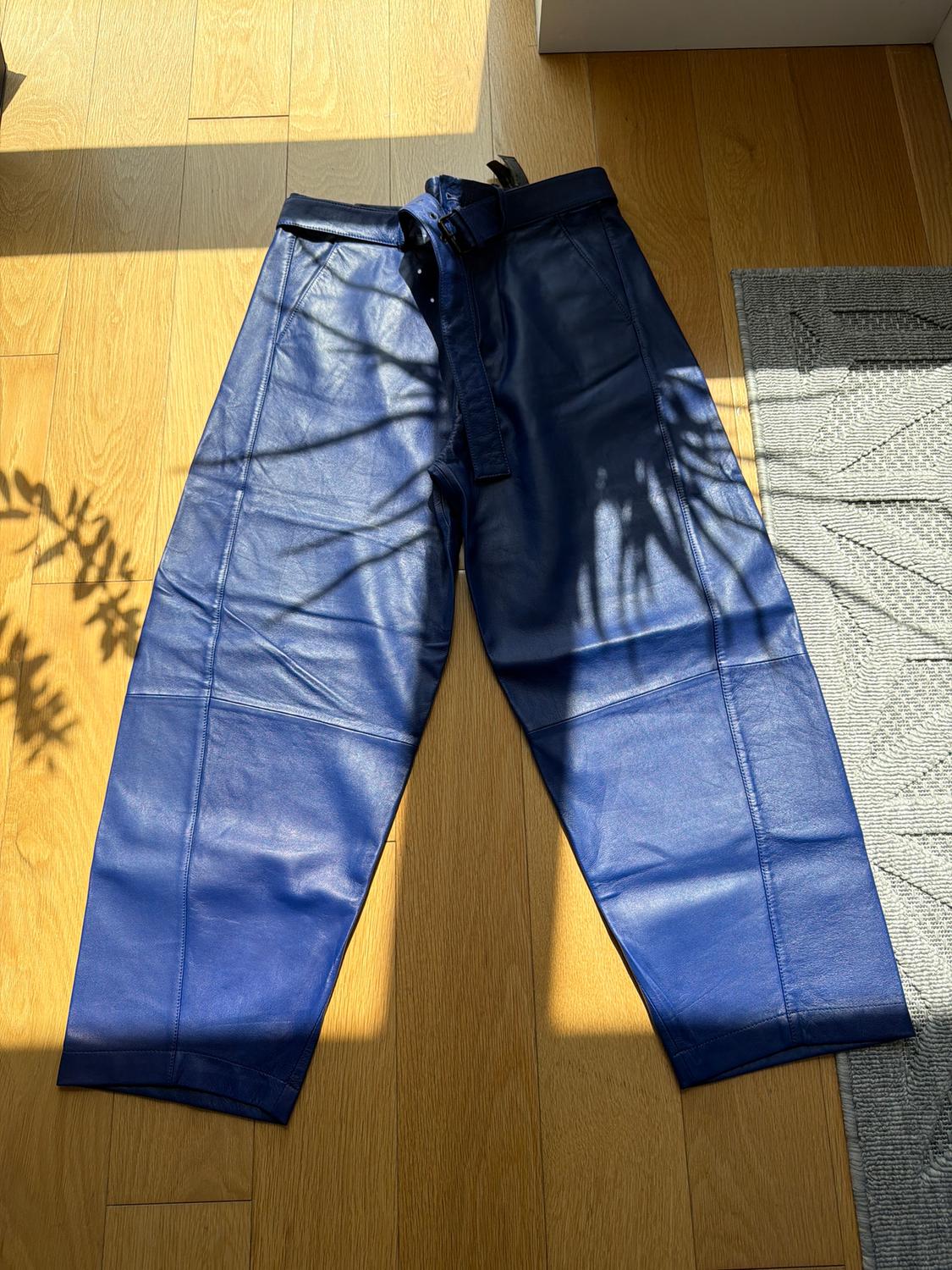 Blue leather pants with a belt, placed on a wooden floor, illuminated by natural light with leaf shadows.