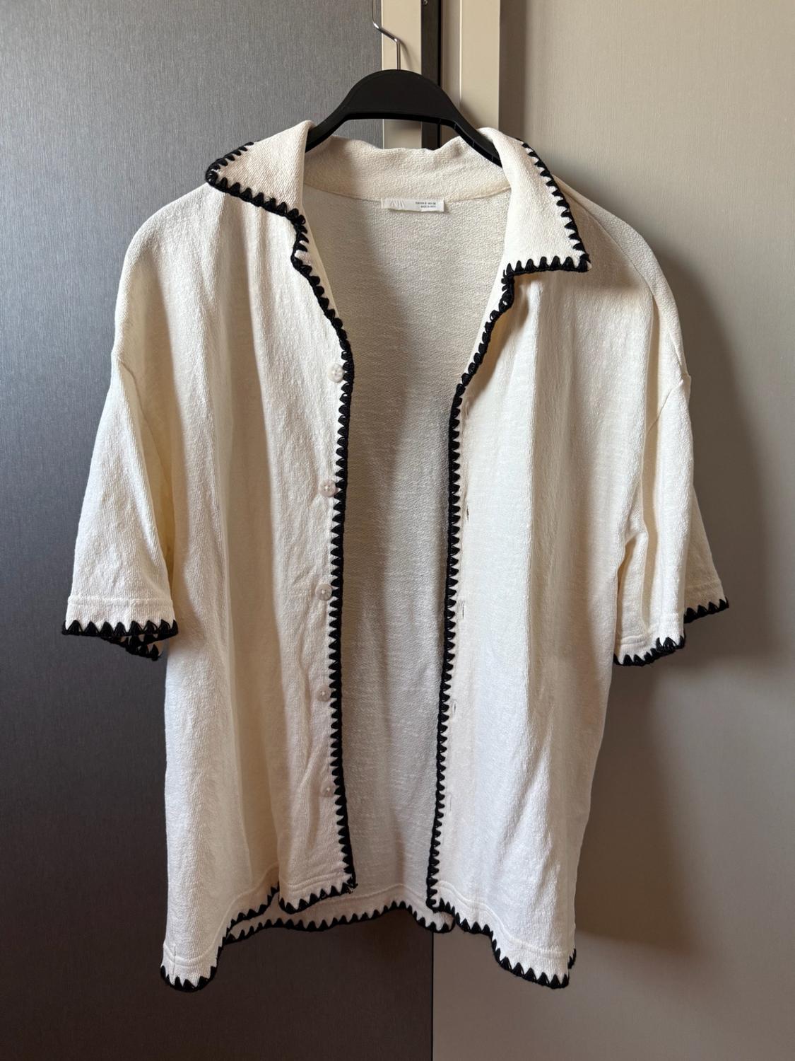 Short-sleeved ecru shirt with black zigzag trim, hanging open on a hanger against a neutral background.