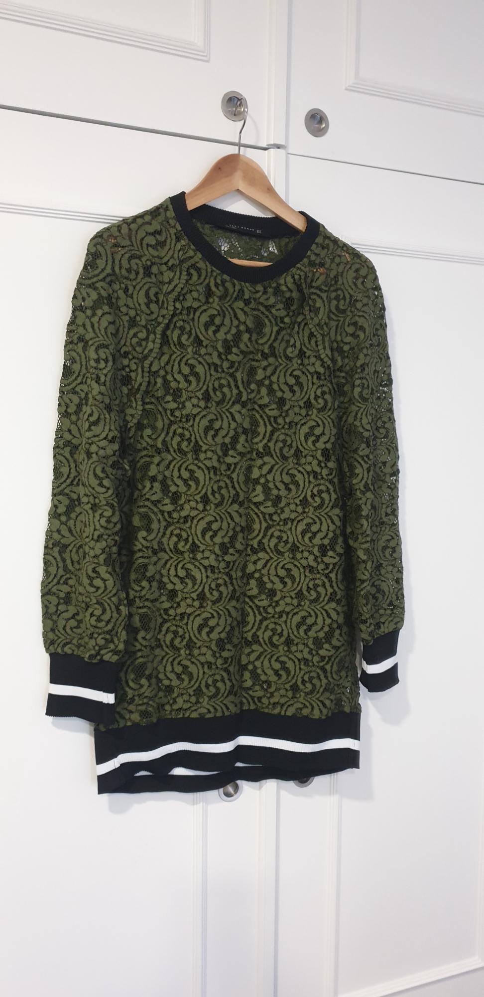 Green lace jumper with floral pattern, black collar and cuffs with white stripes, hanging on a wooden hanger in front of a white wardrobe.