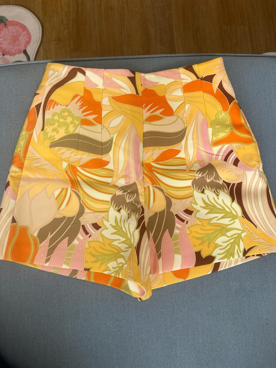 High-waisted shorts with a vibrant retro floral print in orange, yellow and pink tones, on a light blue fabric background.