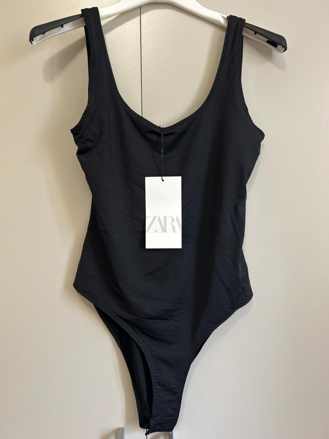 Black strappy body hanging on a hanger. A white label with the text ZARA stands out in the centre. Light background.