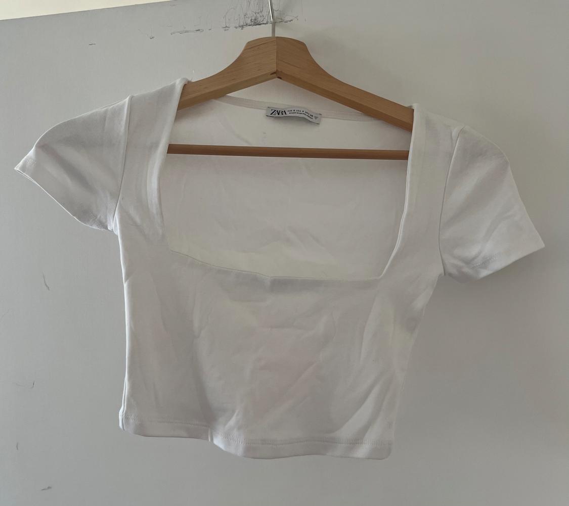 White short-sleeved T-shirt with a square neckline hanging on a wooden hanger against a white wall. Label with the text ZARA.