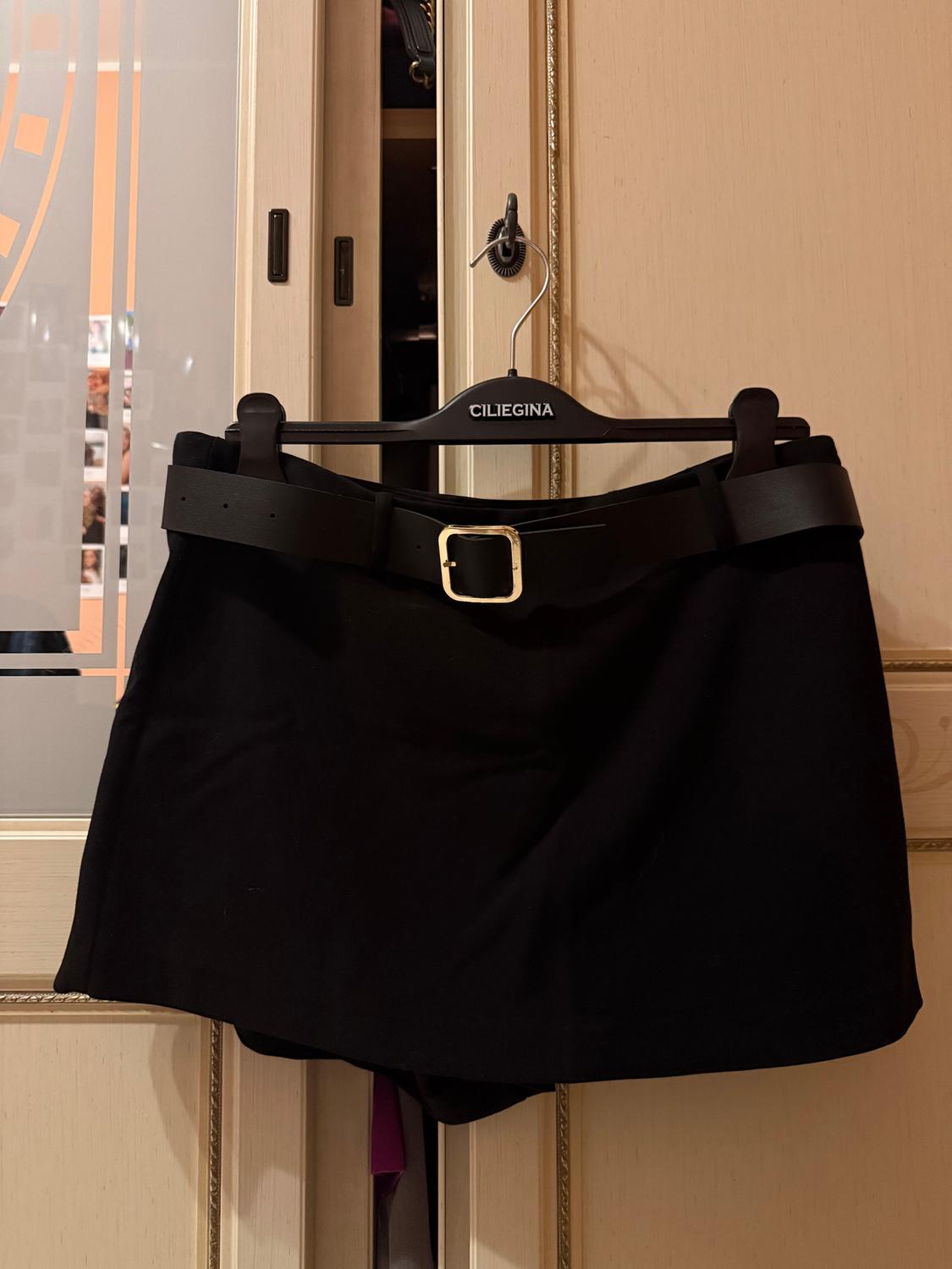 Black skirt with a gold buckle belt hanging on a black hanger with the brand "CILIEGINA" in a light wood wardrobe.