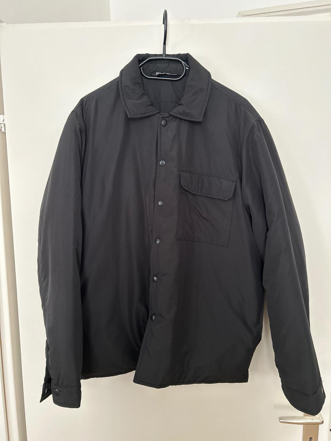 Black long-sleeved shirt hanging on a hanger over a white door. It has front buttons and a chest pocket.
