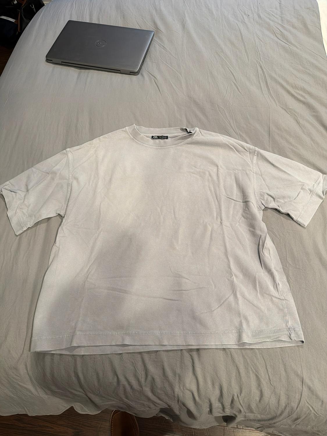 Light grey short-sleeved T-shirt on a bed. Relaxed setting with a closed laptop in the background and the ZARA label visible.