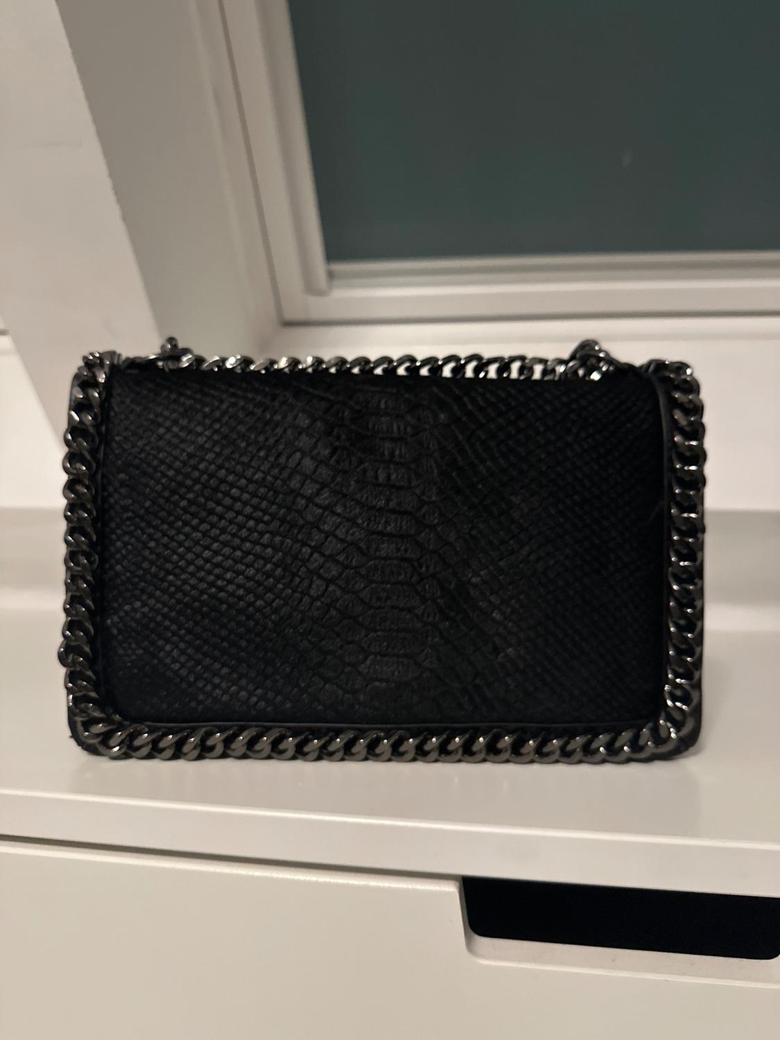 Black handbag with snakeskin texture and metallic chain trim, on a white surface in front of a window.