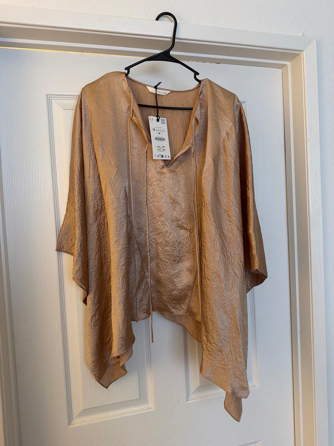 Gold crinkled fabric blouse hanging on a black hanger over a white door. Visible tag with product details.