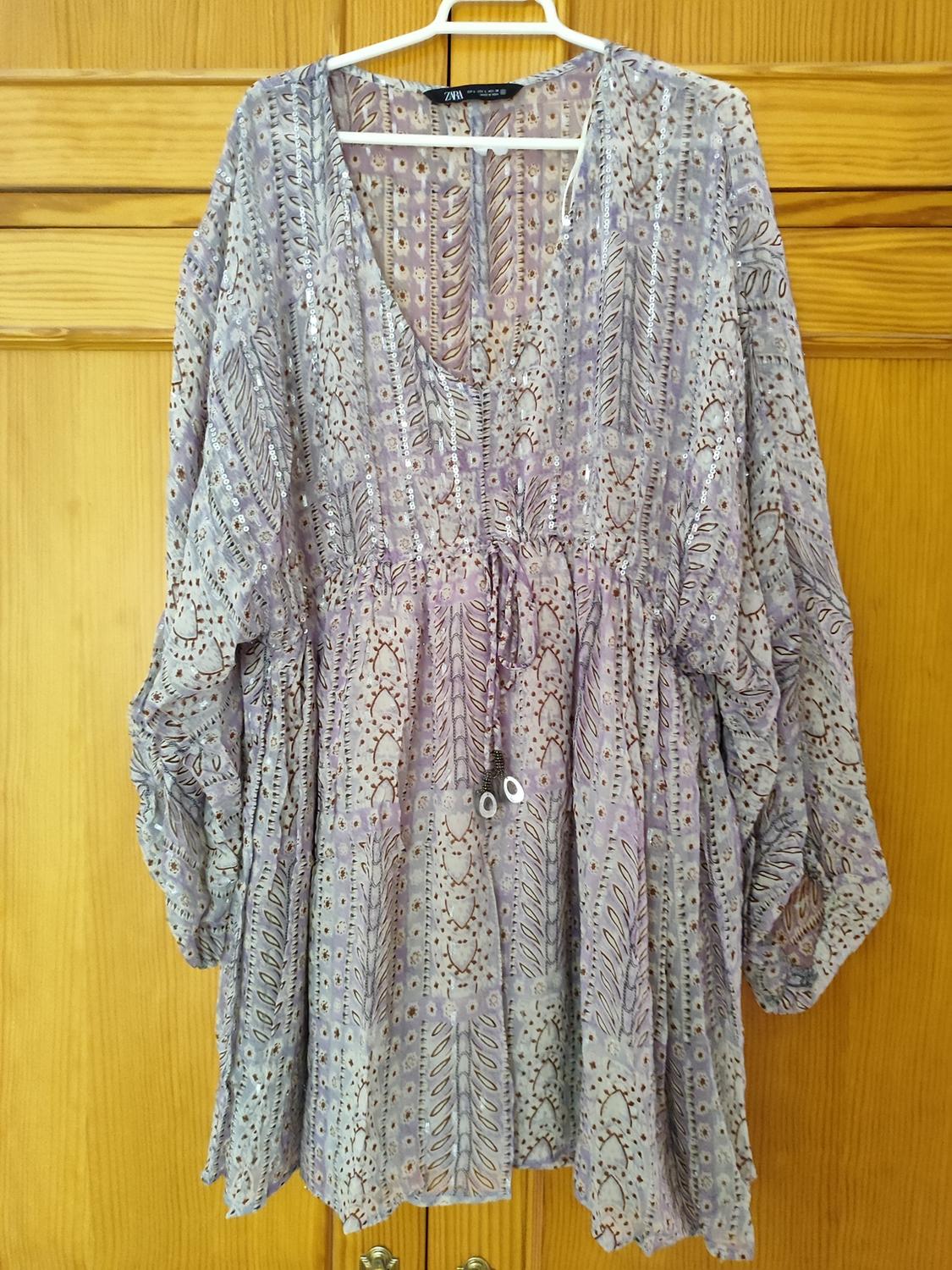 Loose-fitting dress with a geometric print in lilac and beige tones, hanging on a white hanger in front of a wooden door.