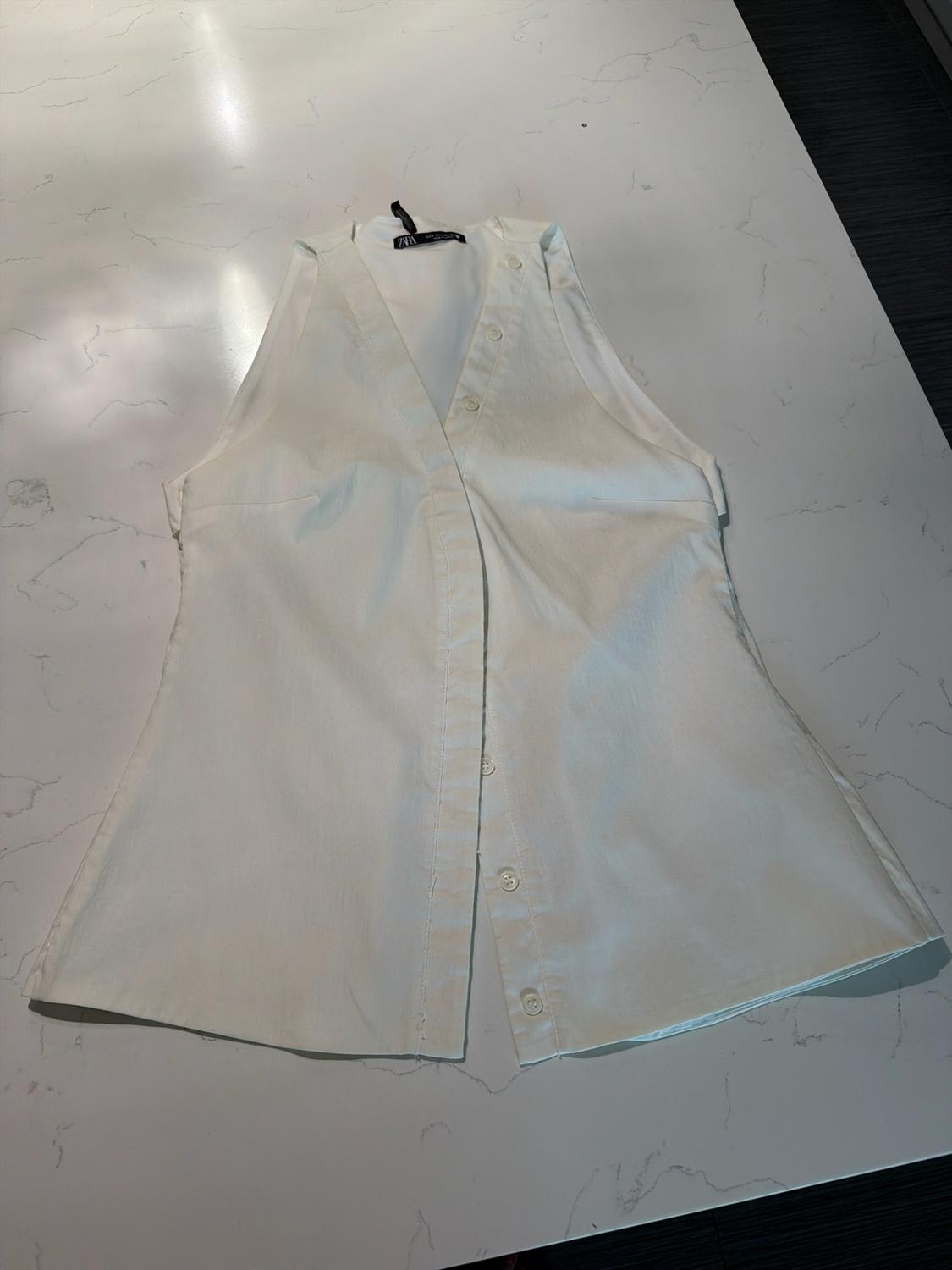 White sleeveless V-neck top with front buttons, on a marble surface. Label with the text ZARA.