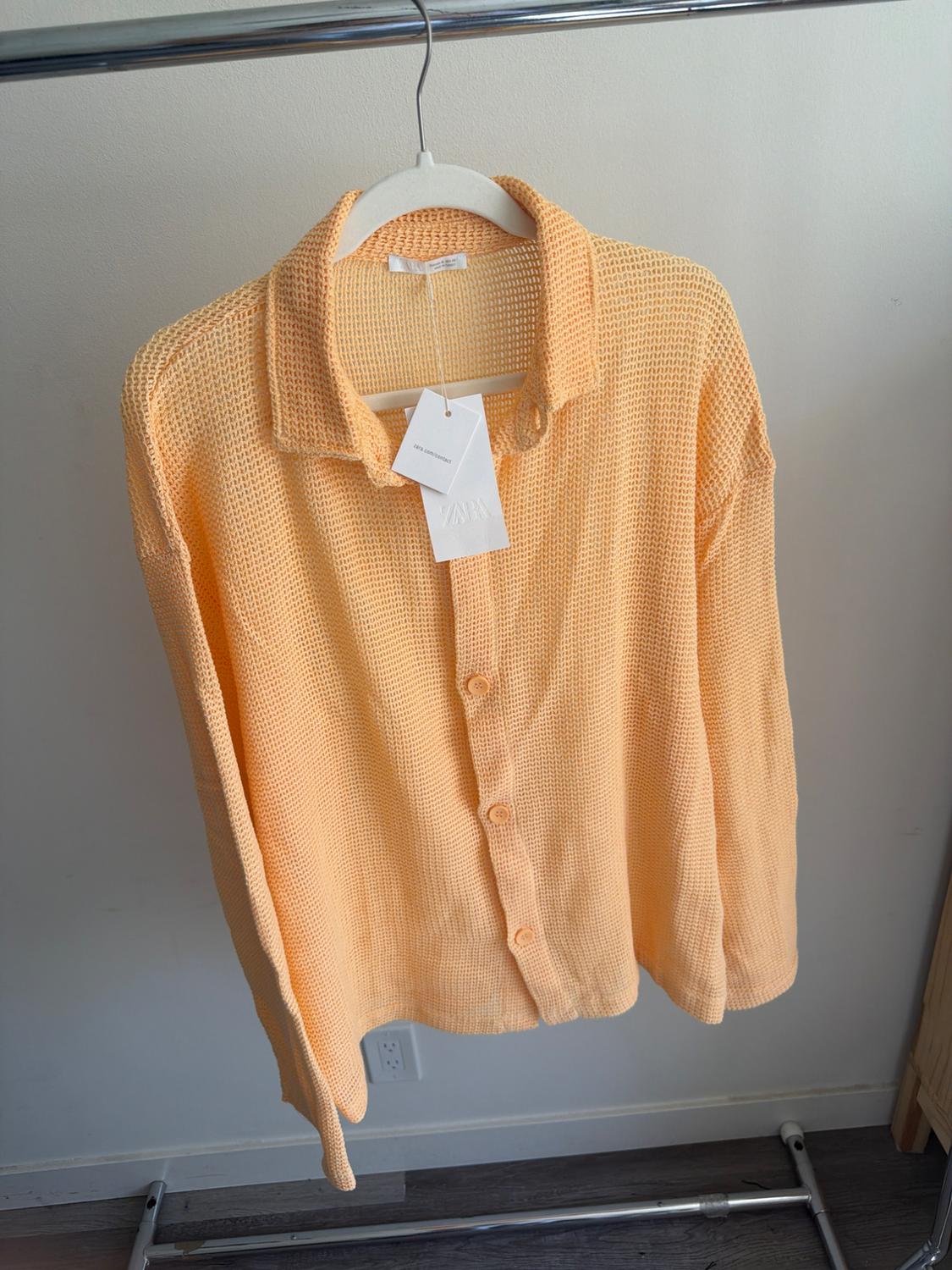 Peach-colored knit shirt hanging on a white hanger, with visible tags. White wall background and wooden floor.