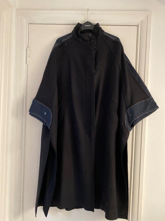 Black cape with denim detailing on the sleeves, hanging on a Topshop hanger over a white door.