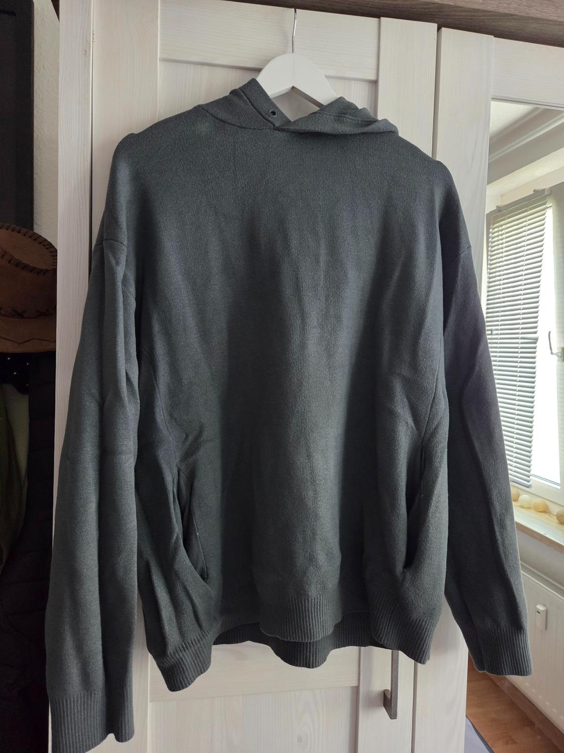 Dark grey knitted hoodie hanging on a white hanger above a light wood wardrobe.