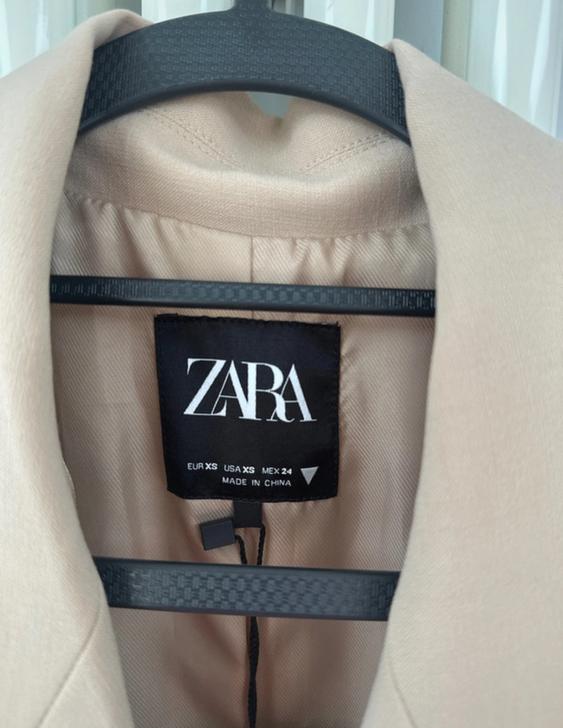 Beige jacket label on black hanger, ZARA brand. Size EUR XS, USA XS, MEX 24. Made in China. White curtain background.