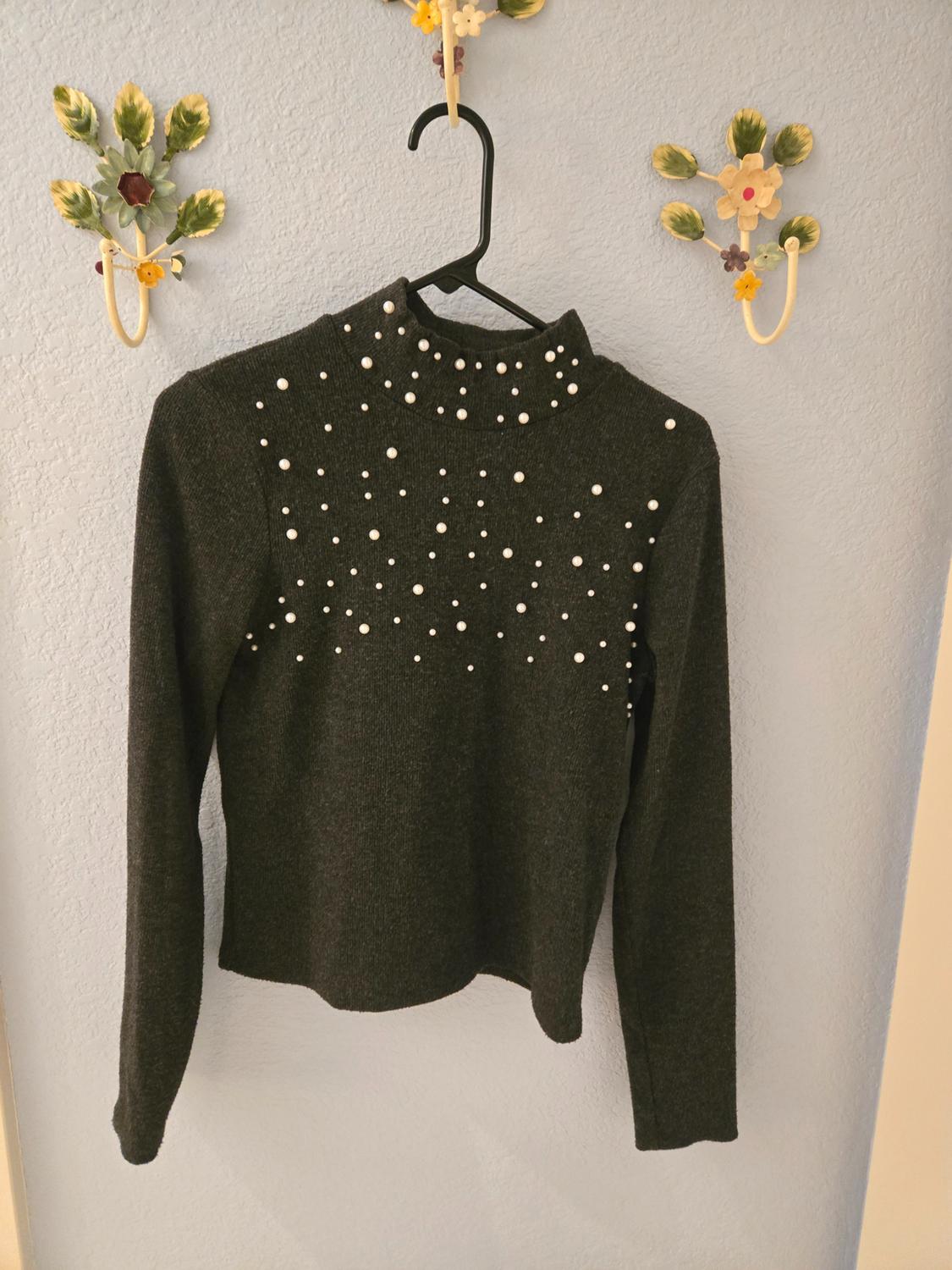 Black polo neck jumper with white pearls on the chest, hanging on a blue wall with decorative floral hooks.