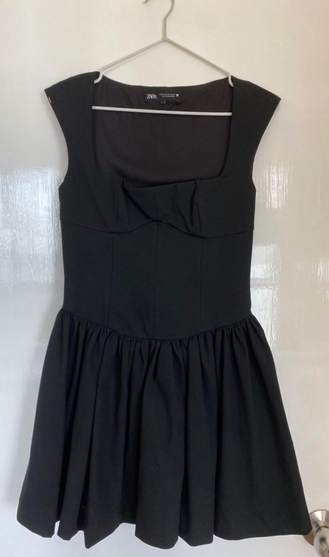 Black sleeveless dress hanging on a white hanger. Square neckline design with a flared skirt. Visible tag: "Zara".