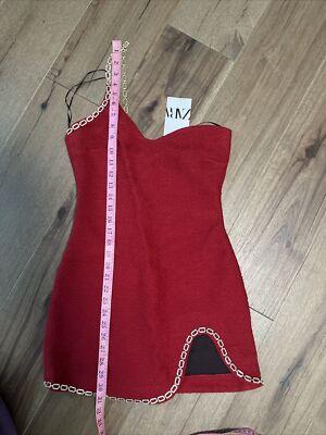 Figure-hugging red dress with thin straps and a gold chain detail on the trim. Pink tape measure on the dress. Wooden background.