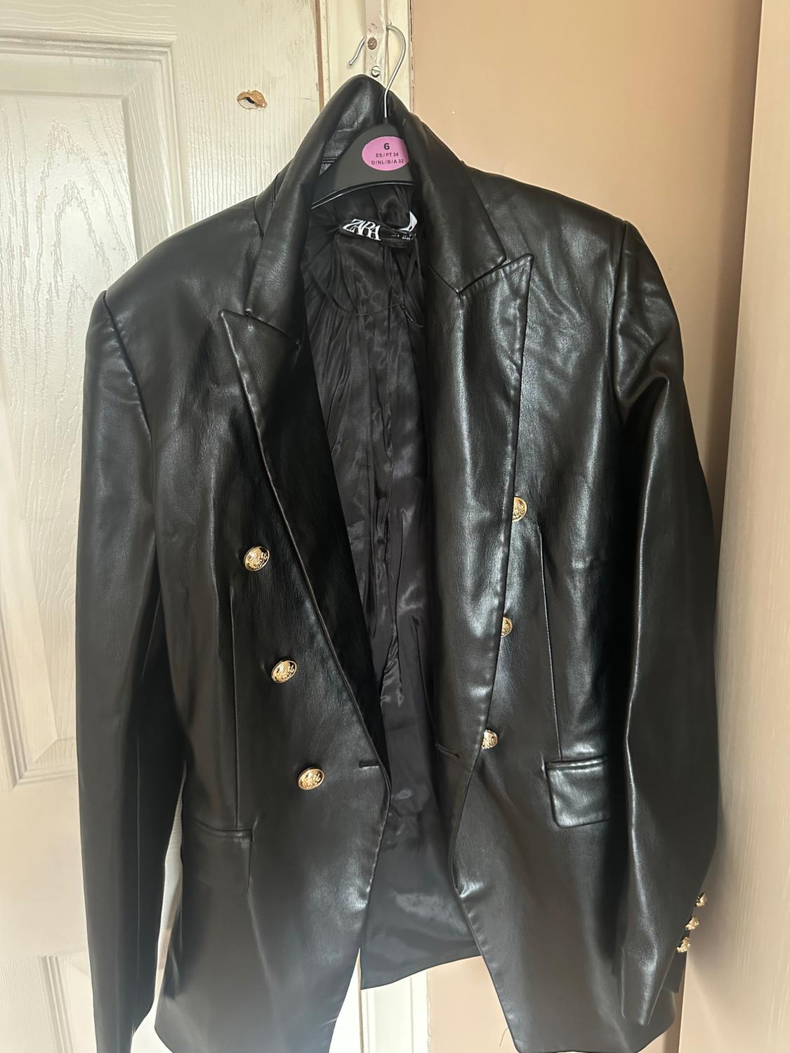 Black jacket hanging on a hanger with gold buttons. White door background and beige wall. Visible tag on the hanger.