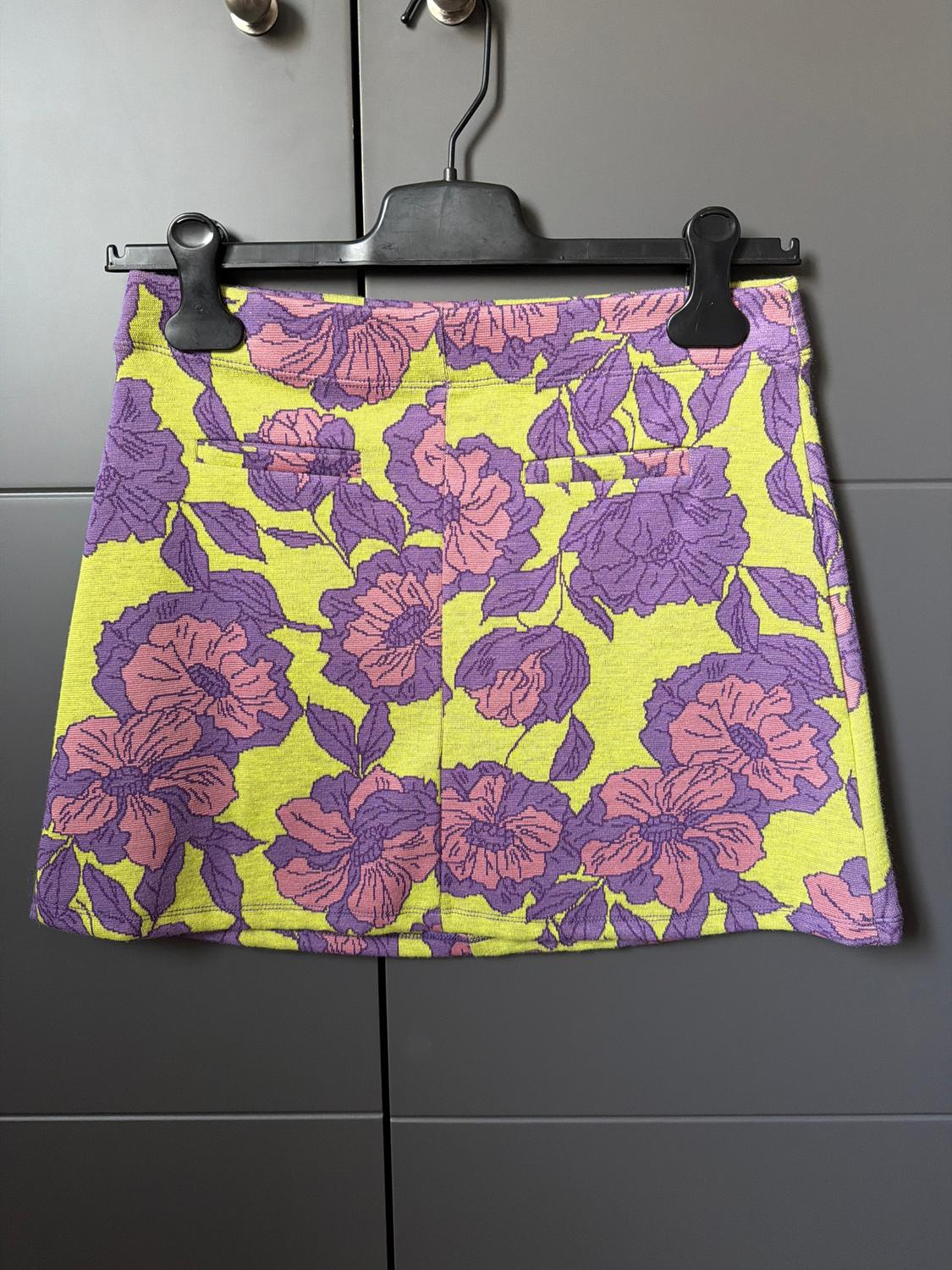 Mini skirt with floral print in purple and pink tones on a yellow background, hanging on a black hanger in front of a grey wardrobe.